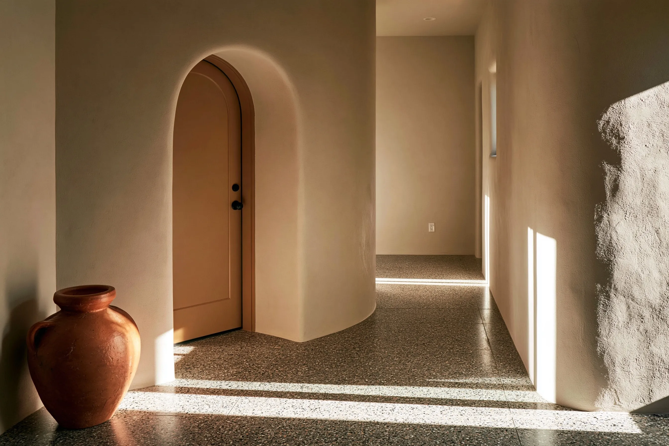 Entry hall and corridor with a salt-and-pepper fine-aggregate polished floor, arched stucco doorway, terracotta pot, daylight, low angle